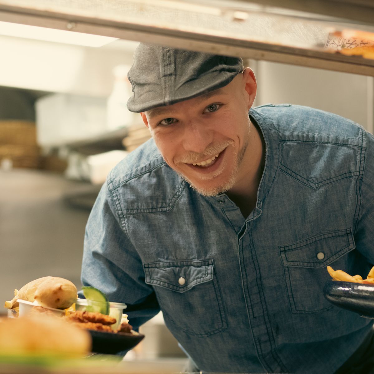 Chef souriant en chemise en denim servant des repas fraîchement cuisinés depuis la cuisine